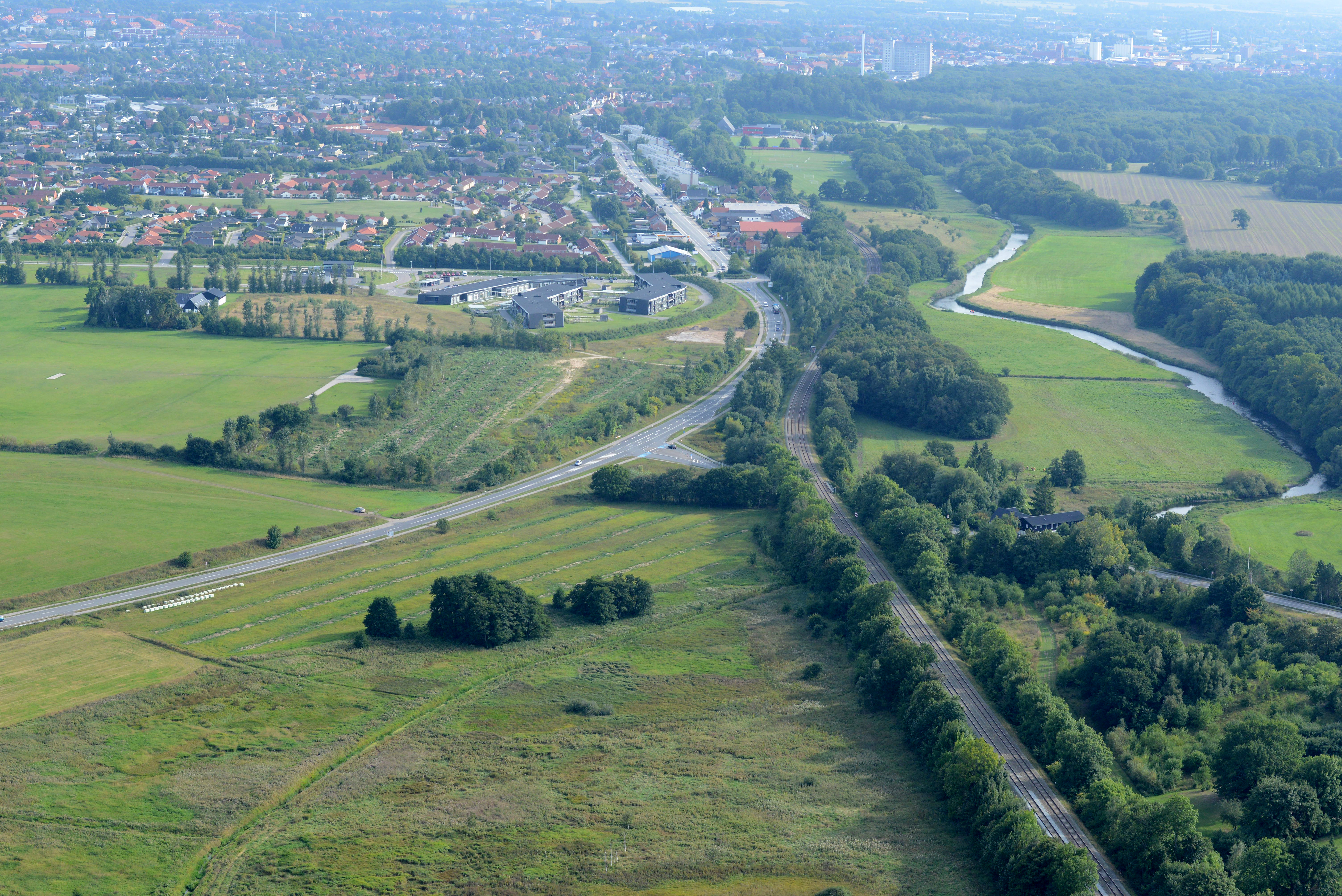 Luftfoto af et landskab med en mindre by i baggrunden og en vej og banestrækning, der løber gennem landskabet