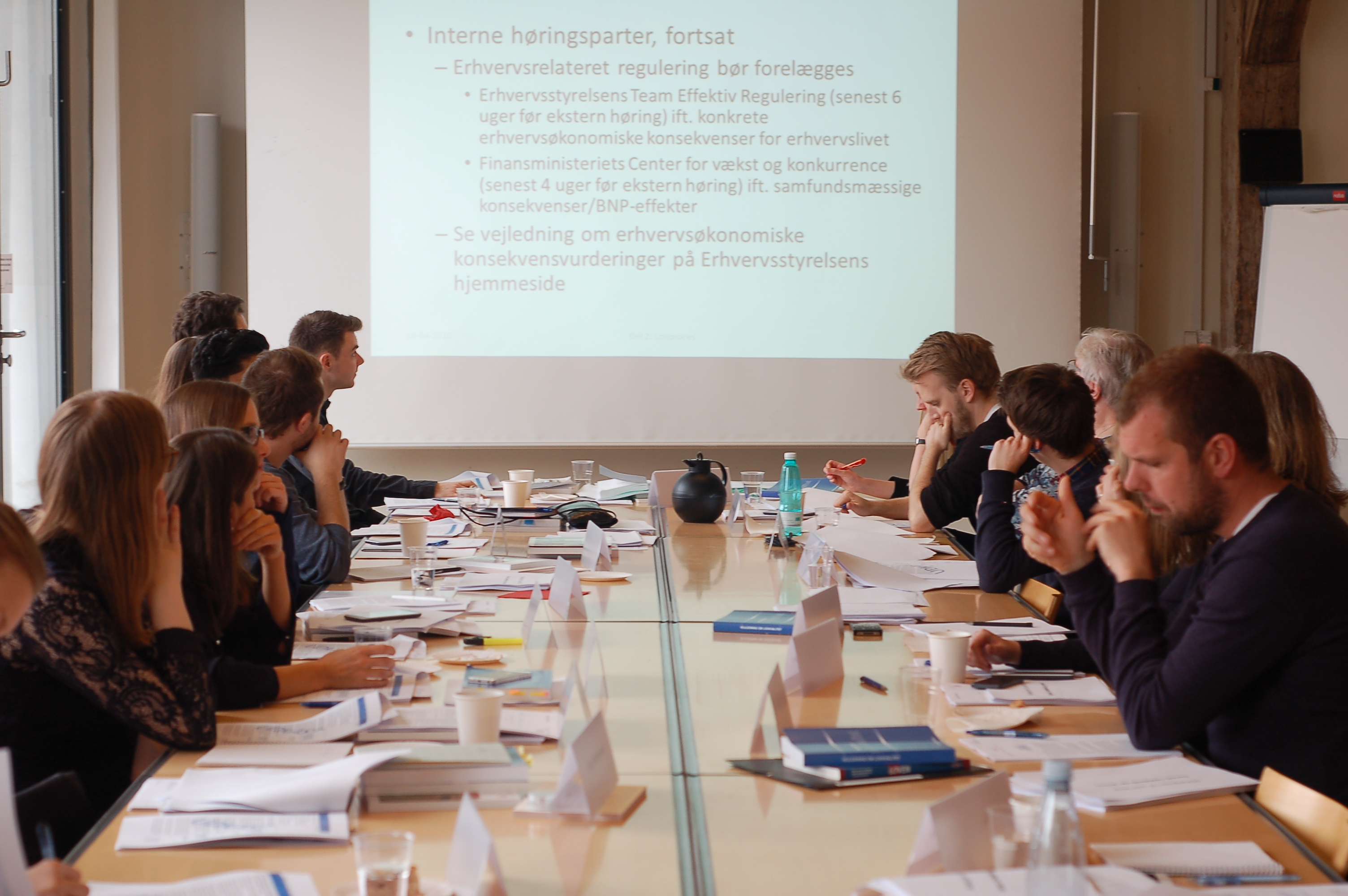 En flok mennesker sidder ved et stort mødebord med deres papirer og en skærm for enden af lokalet.