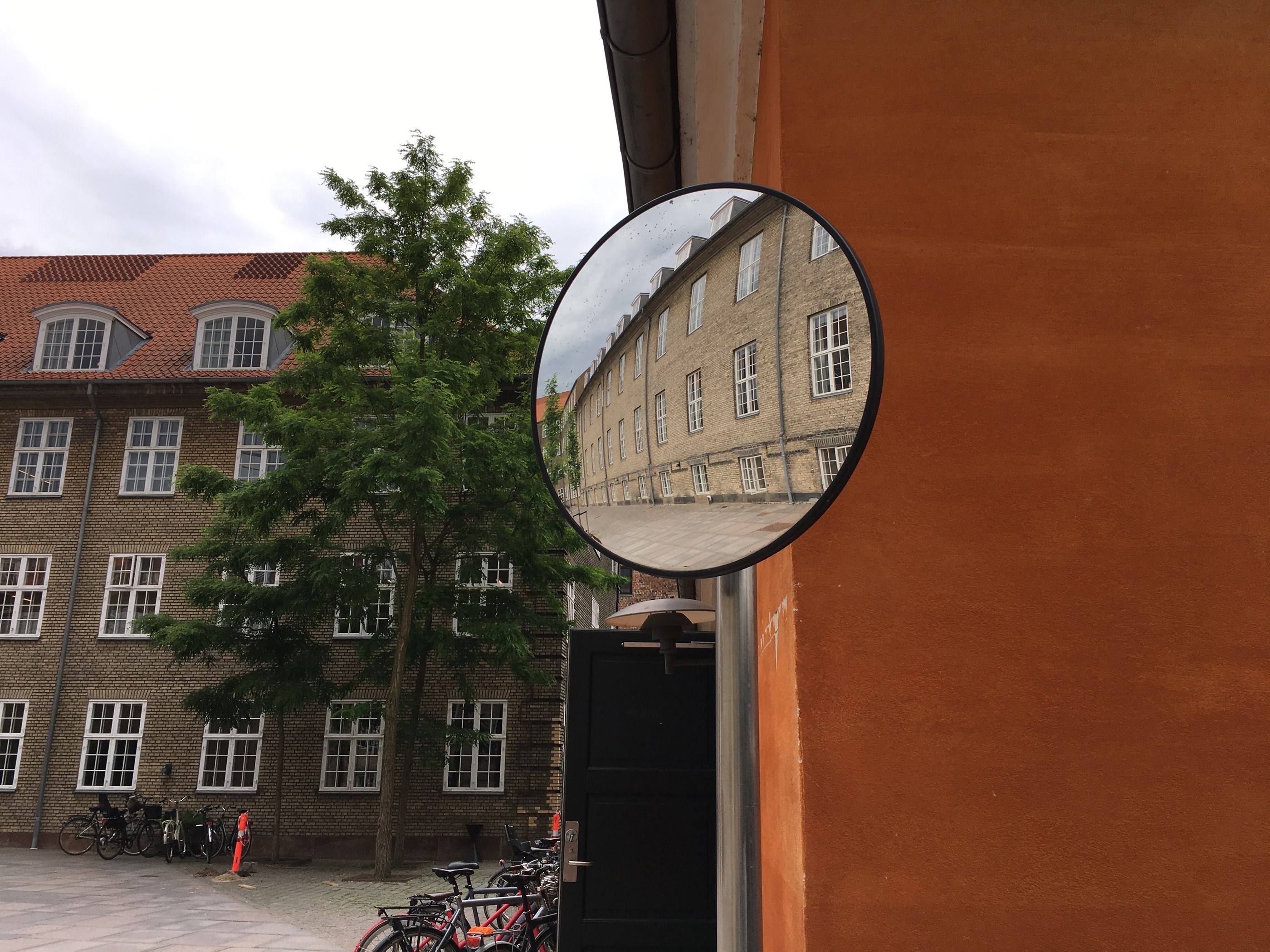 På midten af billedet ses et trafiksepejl. Dette genspejler en murstensbygning i tre etager. I baggrunden ses en lignende bygning, et træ og en orange mur.