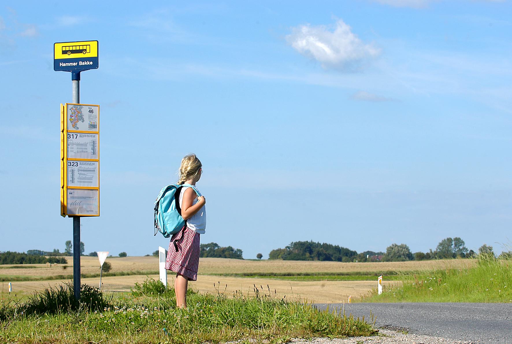 Pige står ved et busstoppested i landlige omgivelser.