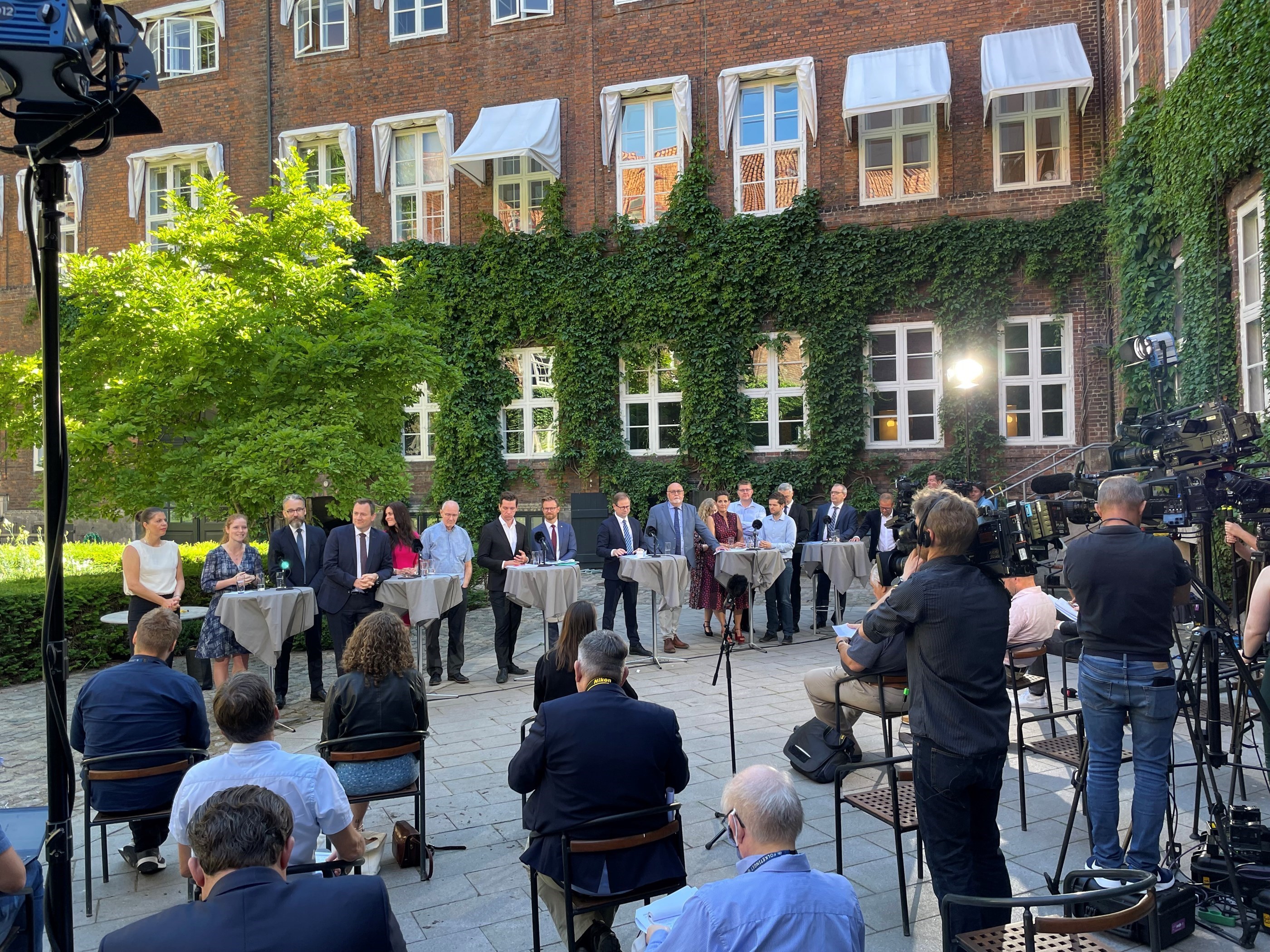 Foran en mur med grønne planter står en række mennsker ved borde. I forgrunden ses et kamera der filmer disse og ni personer der sidder med ryggen til os.