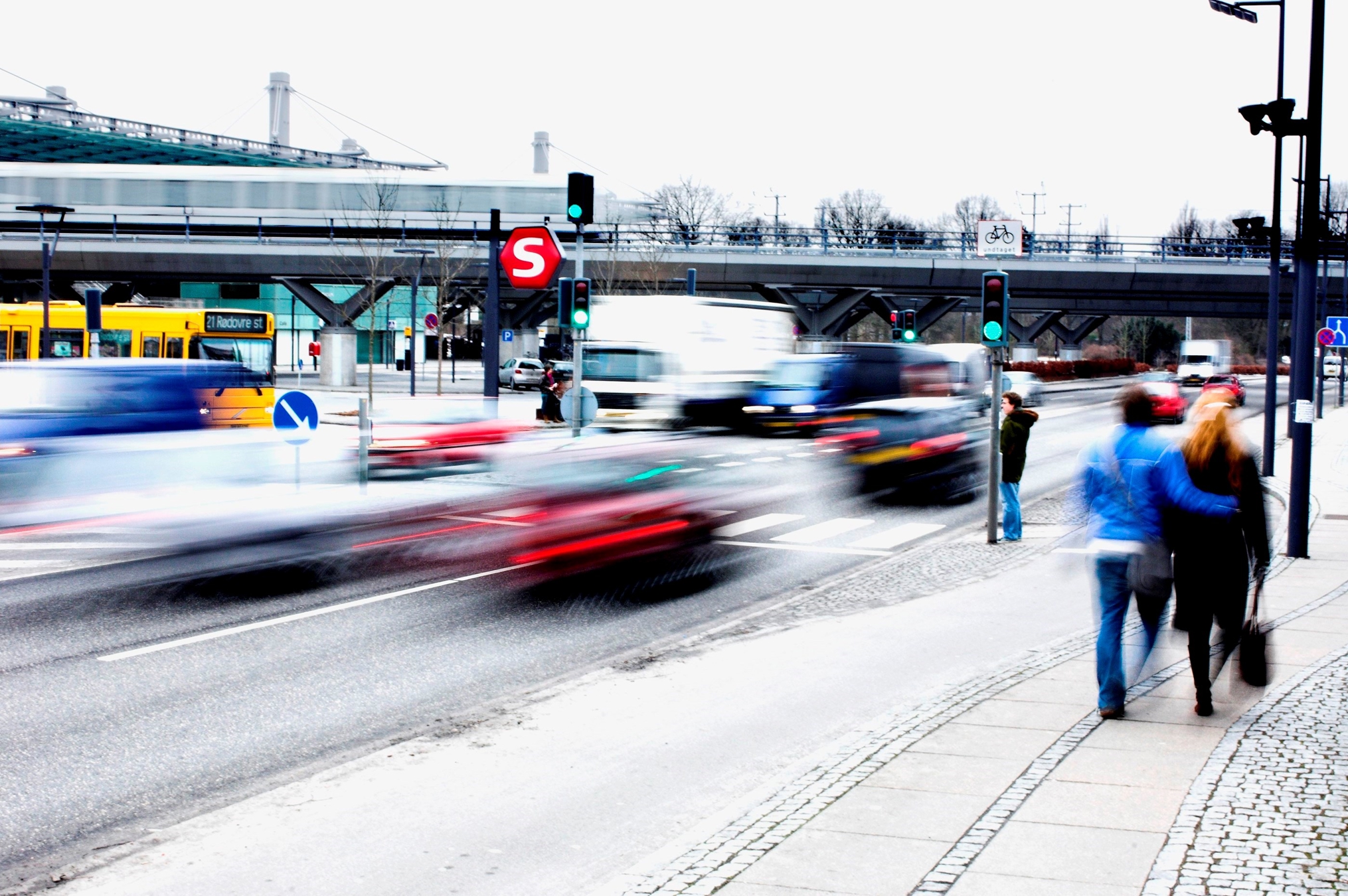 Et lyskryds, hvor bilerne og fodgængerne er slørede. I baggrunden ses et s-togs skilt.